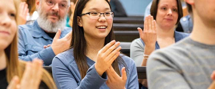 A diverse group of students in an ASL class at FLCC, with their hands raised in a lecture hall.