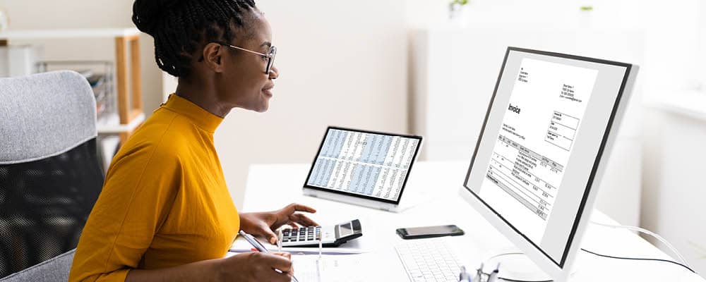 An accountant sits at her desk reviewing invoices on a screen while inputting figures into a calculator.